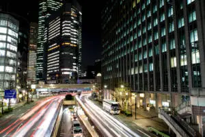 Busy Tokyo city street at night, with light trails from passing cars and tall buildings glowing in the background. Photo by Tia Haygood.