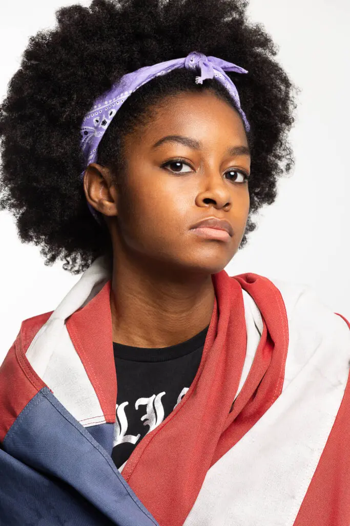 A black young woman with curly hair, and da country's flag draped over her dress, facing the camera - Headshot photos