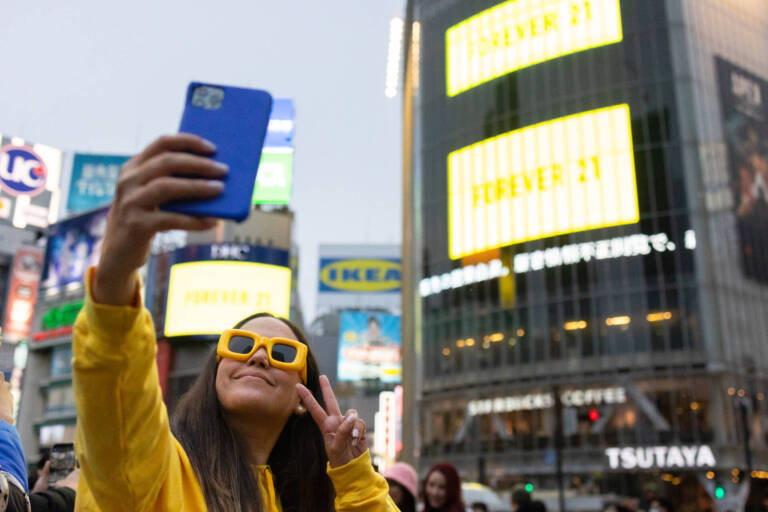 A woman in a yellow dress and yellow-framed glasses taking a selfie with a building in Tokyo - Tokyo Portrait Photographer