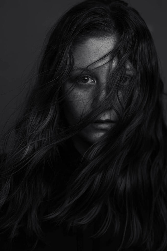 A black and white photo of a young woman with long hair covering her face by the wind. 